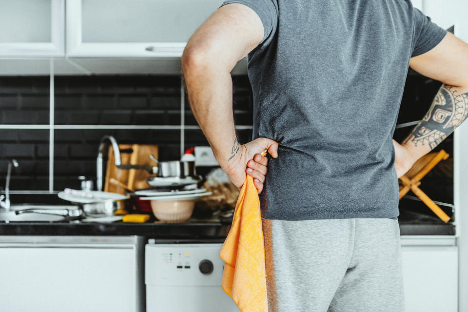 A man with a tattooed arm in casual attire stands with his back to the camera in a kitchen with unwashed dishes in the sink and on the counter