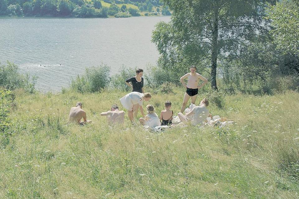 Characters wear 1940s-era bathing suits in an appealing green field on the banks of a river.
