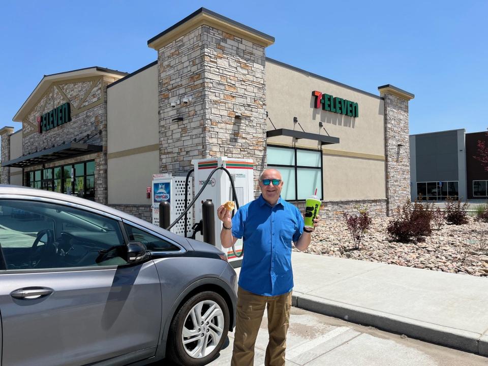 Nigel Zeid holding up snacks while standing next to an electric vehicle that is charging in front of a 7 Eleven.