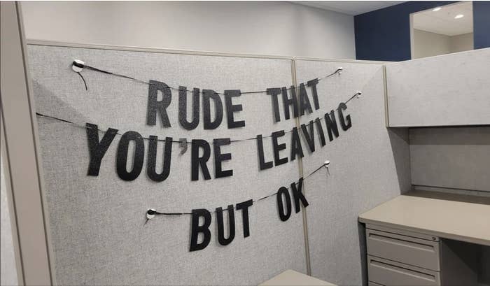 Banner across office cubicle reads "RUDE THAT YOU'RE LEAVING BUT OK", possibly a coworker's humorous farewell