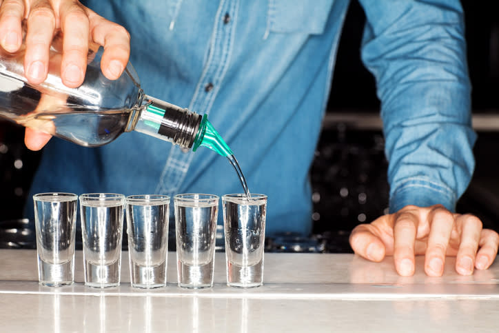 Bartender pouring clear liquid into a row of shot glasses on a bar counter