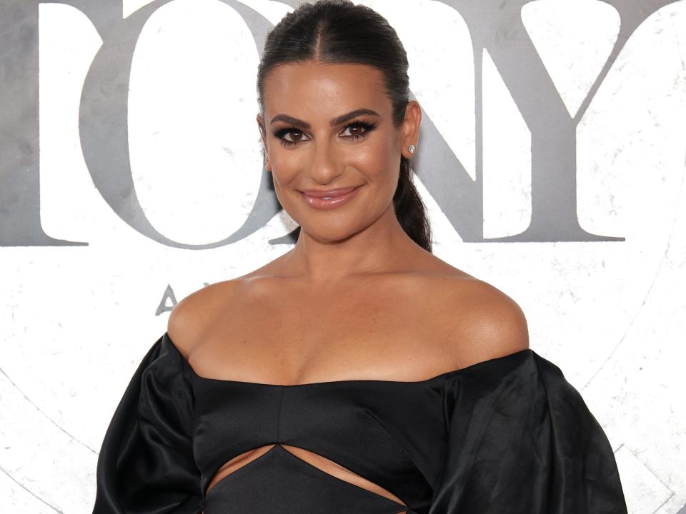 Lea Michele, in an off-the-shoulder black gown with cut-outs, smiles while posing on the red carpet at the 2022 Tony Awards.