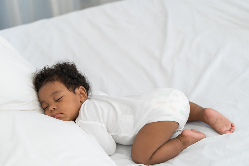 A baby sleeps peacefully on a bed, lying face down with legs tucked in, wearing a white onesie