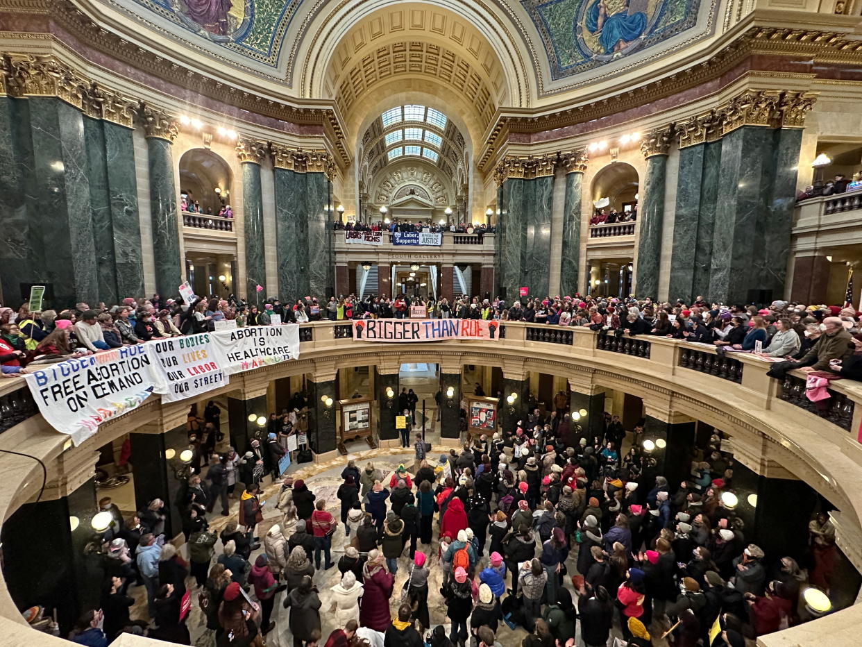 Demonstrators, many in pink wool hats, gather under an ornate cupola at the State Capitol, with banners over an upper balcony reading Bigger than Roe and Abortion Is Healthcare.