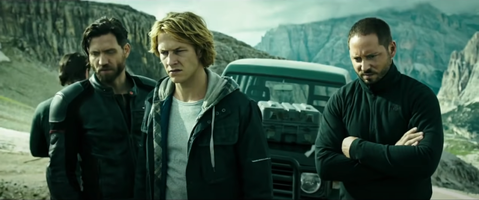 A group of men stand in front of a car atop a cliff, looking seriously at something