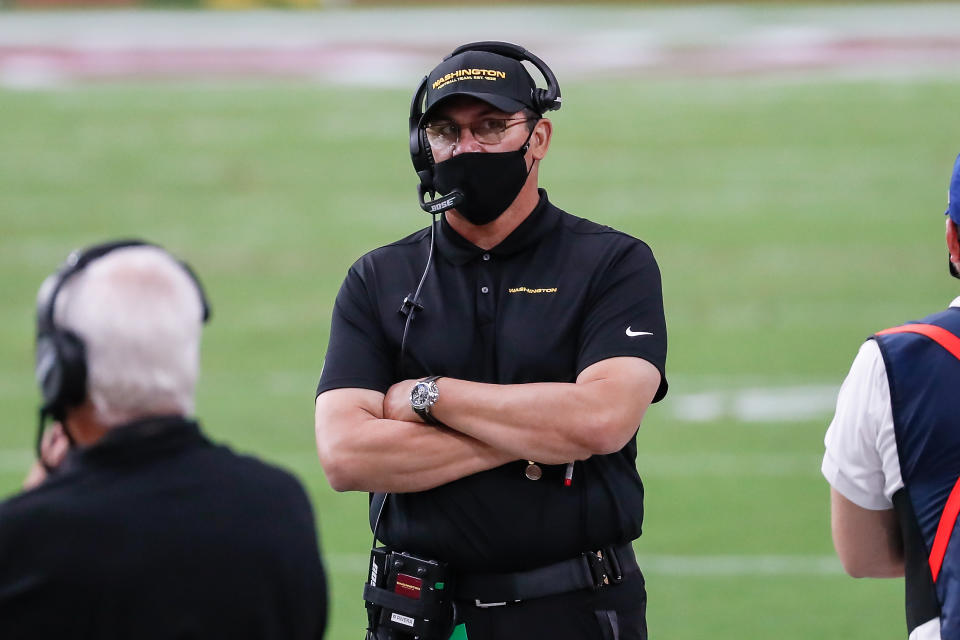 Ron Rivera in a black mask stands on a field, arms crossed. 