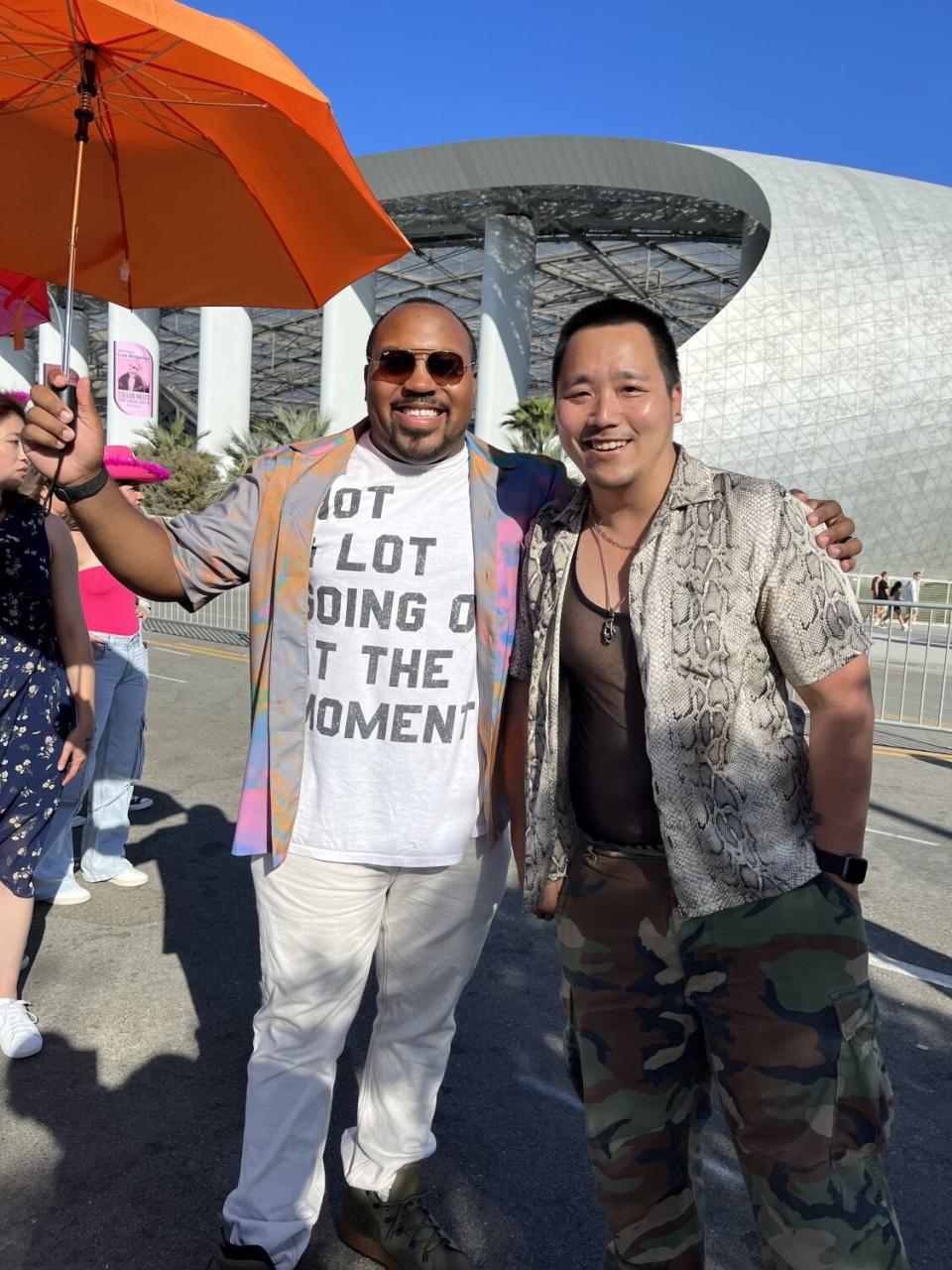 Two smiling men pose together holding an orange umbrella outside a concert venue.