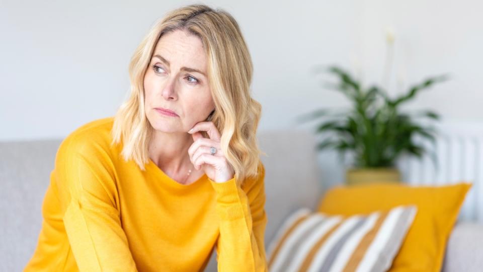 A blonde woman in a yellow top looking out the window because she feels sad and tired due to allergies