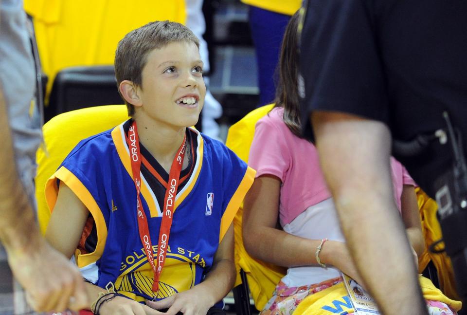 aaron hern wearing a blue basketball jersey, sitting in seats at a basketball court, smiling and looking up at someone off camera