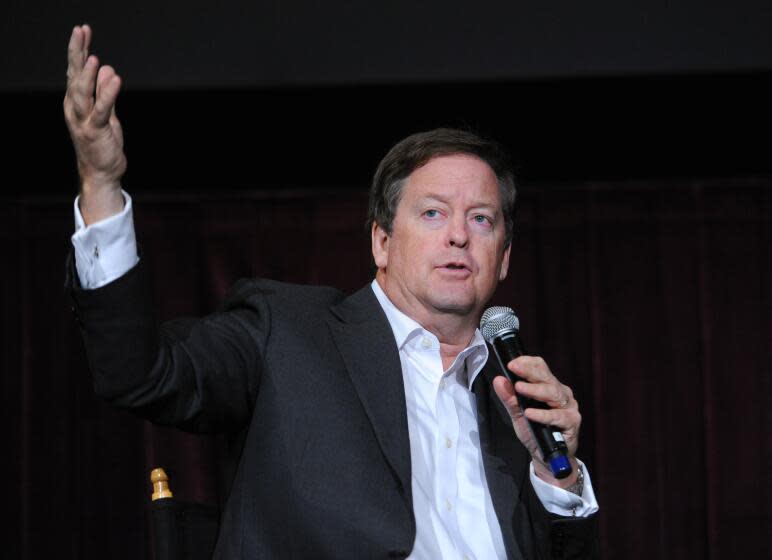 A man in a suit jacket and light-colored dress shirt speaking into a microphone while raising his right arm