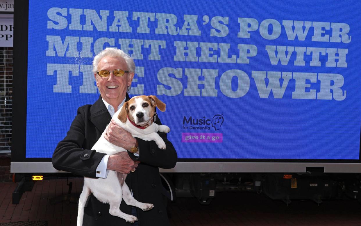Tony Christie holds up his dog in front of a poster by Music for Dementia