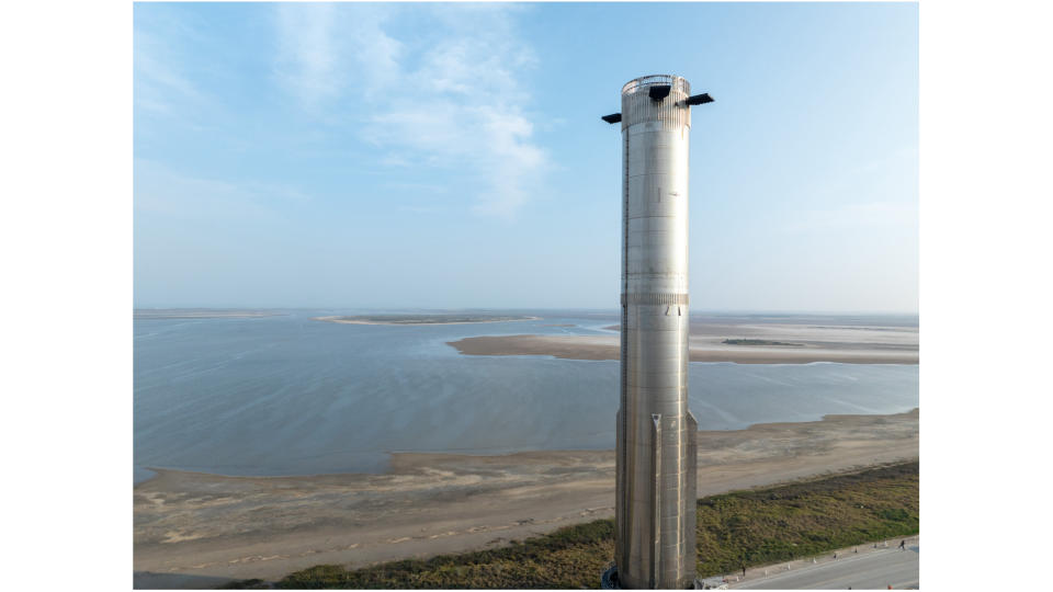 a very tall, silver rocket rolls along on a truck on a road next to the ocean