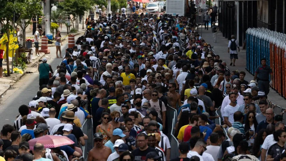 Fotografia colorida mostra multidão em fila em frente a estádio