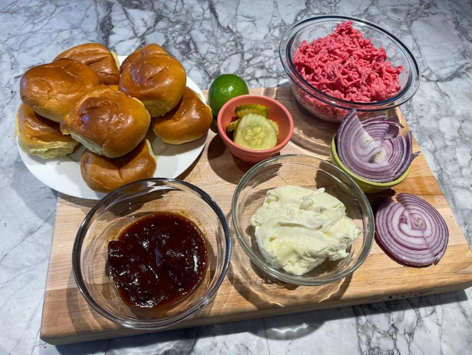 A wooden cutting board filled with hamburger buns, barbecue sauce, mayo, sliced pickles, slices of red onions, a bowl of ground beef, and a lime