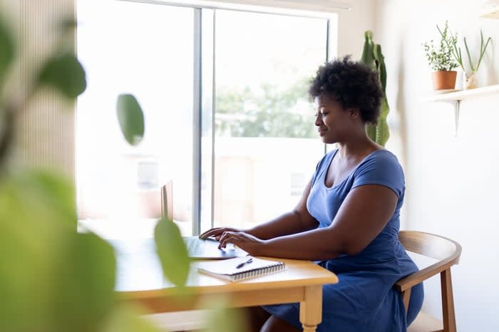 A person sitting at a desk in a sunny home office and typing on a laptop.