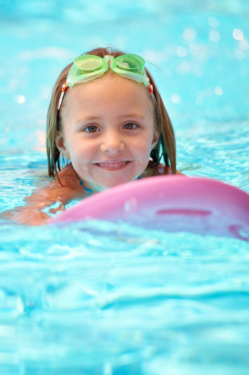 A young child wearing swim goggles smiles while holding a pink kickboard in a swimming pool