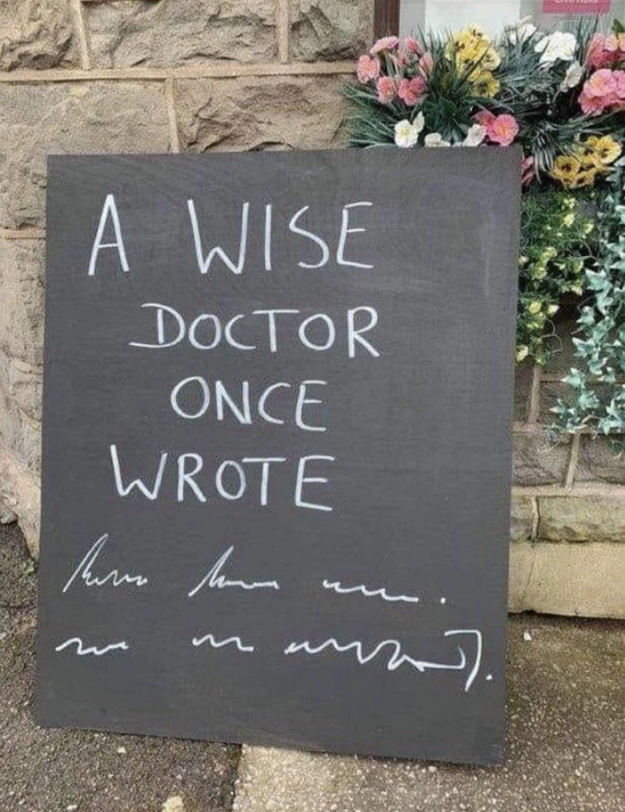 A handwritten chalkboard sign reads, "A wise doctor once wrote" followed by an illegible scribble, placed outside a stone building with flowers