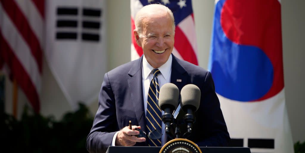 joe biden, wearing a dark blue suit jacket, white shirt, and striped tie, smiles while standing at a podium with two microphones on it, with american and south korean flags behind him