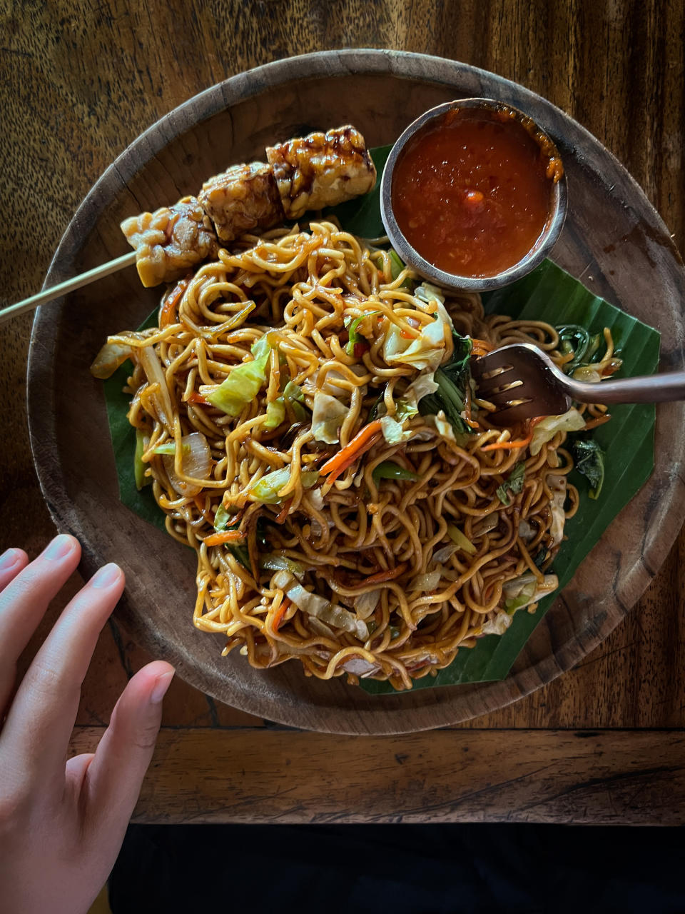 A plate of noodles with vegetables and meat on skewers, with sauce on the side, the hand is visible