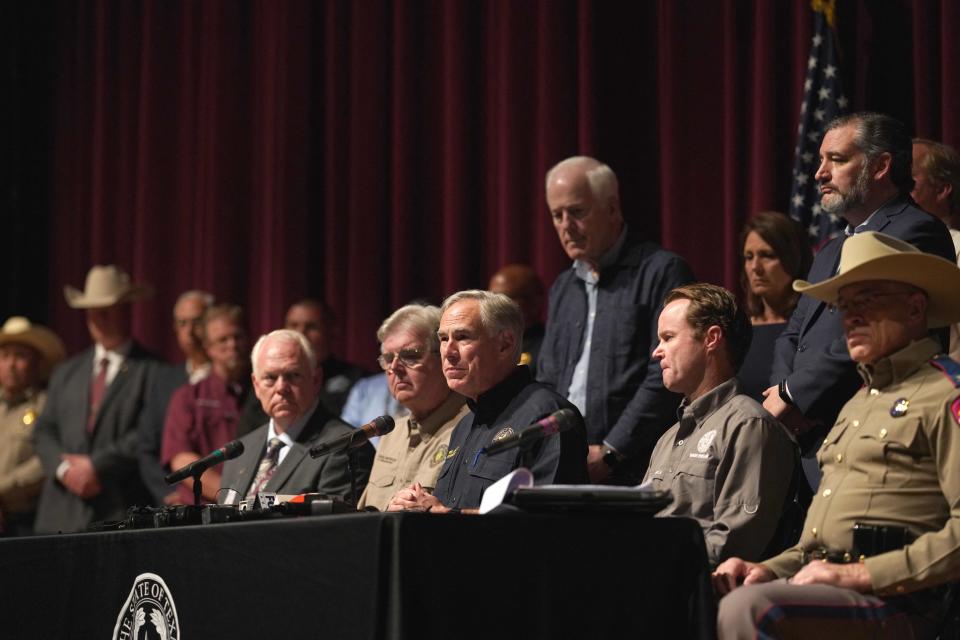 Greg Abbott and a dozen or so other people are on stage behind a table with microphones in front of a red curtain and American flag.