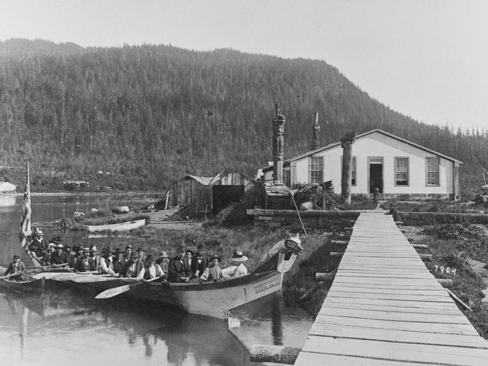 Totem poles visible in the grounds of Chief Shakes house as a Tlingit canoe - named 'Brown Bear' - carrying a funeral party arrives at Fort Wrangel on Wrangell Island in the Alexander Archipelago, Alaska, 1890. Chief Shakes is a leadership title bestowed by the Tlingit, the northernmost peoples of the northwest coast of North America, residing on the islands and coastal lands of southern Alaska.