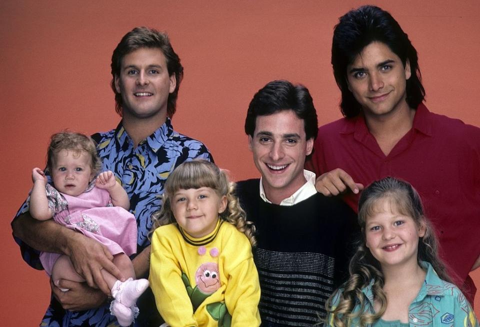 Cast of "Full House" with three men standing and three young girls, two sitting and one held, in casual clothes