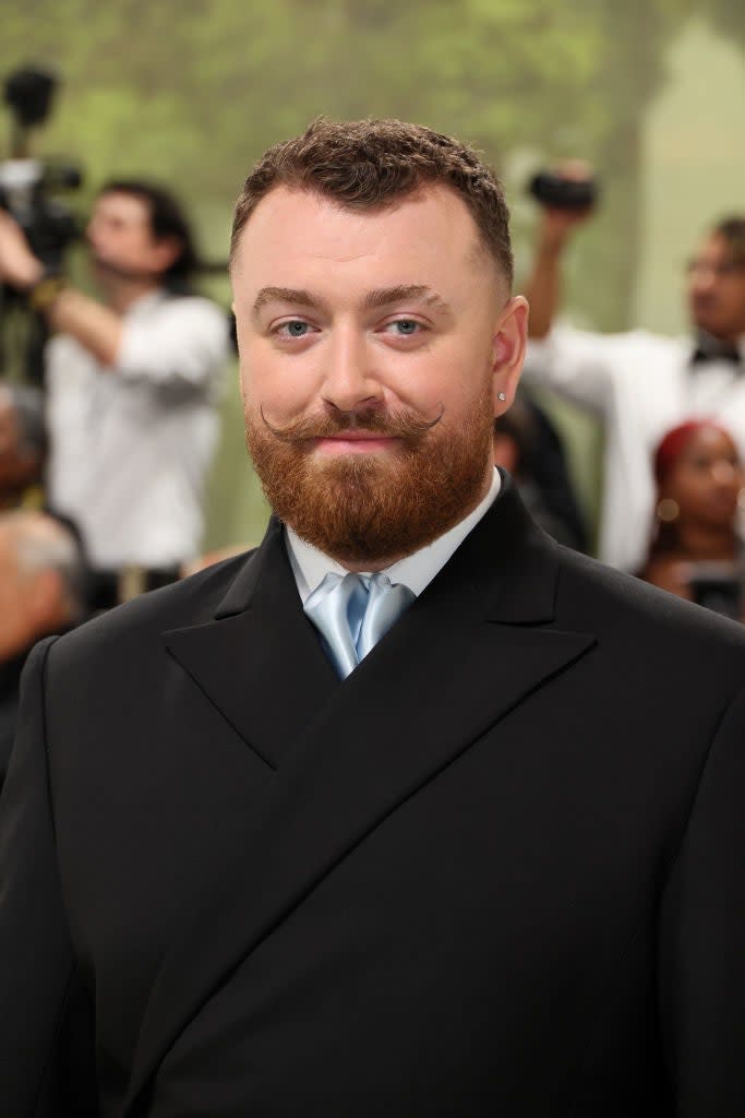 Sam Smith on the red carpet, wearing a classic black suit with a blue tie, sporting a well-groomed beard and mustache