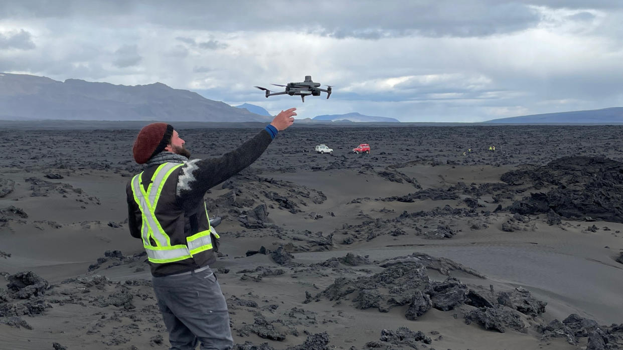  a drone alights on the hand of a man standing in the middle of a black lava field with mountains in the backgroud 