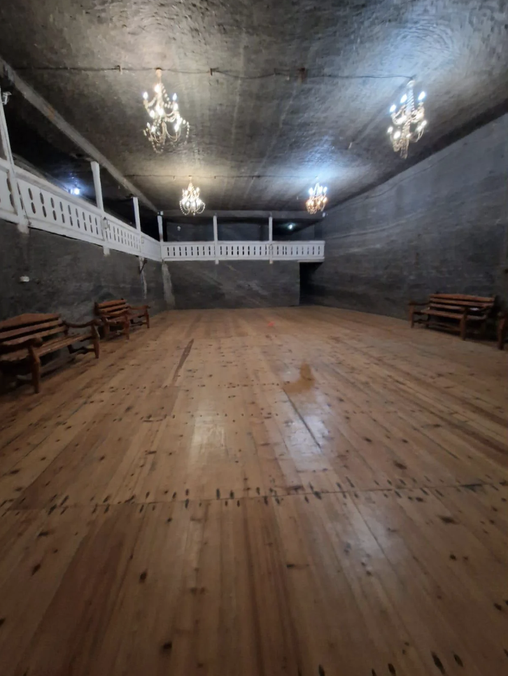 Interior of a dimly lit historic hall with wooden floors, benches, and chandeliers