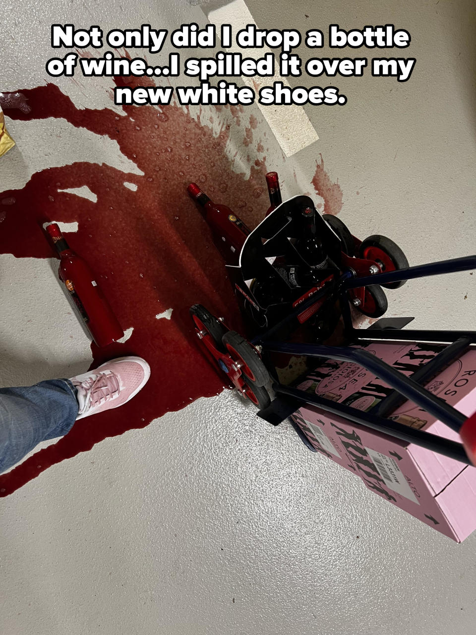 Overturned cart with spilled red wine on a garage floor, next to a person's foot wearing a stained sneaker, with caption: "Not only did I drop a bottle of wine, I spilled it over my new white shoes"