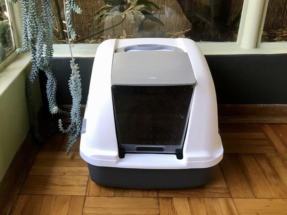 A white and black hooded Catit Jumbo Litter Pan on wooden floor in a corner of a room.