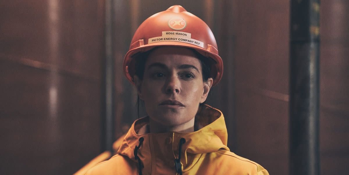 emily hampshire in the rig, wearing a hard hat and hi vis and looking glum