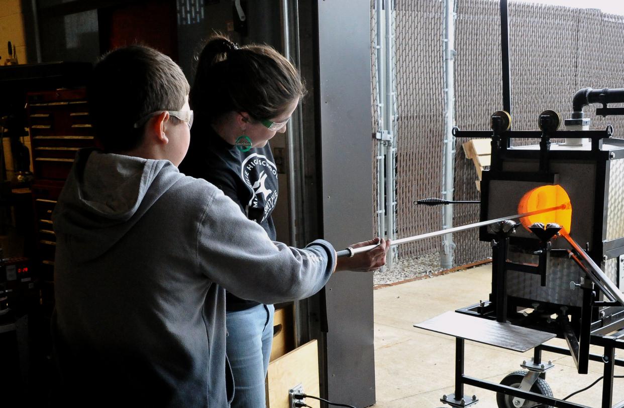 Clayton Saurer holds a hot piece of glass on a rod in the blast furnace as Smithville art teacher jennifer Winkler helps him to make a flower..