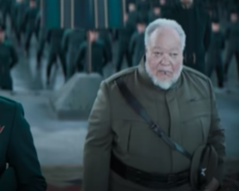 Stephen wears an olive-green military uniform and looks off-camera in a "Dune" still, in the background are hundreds of more soldiers