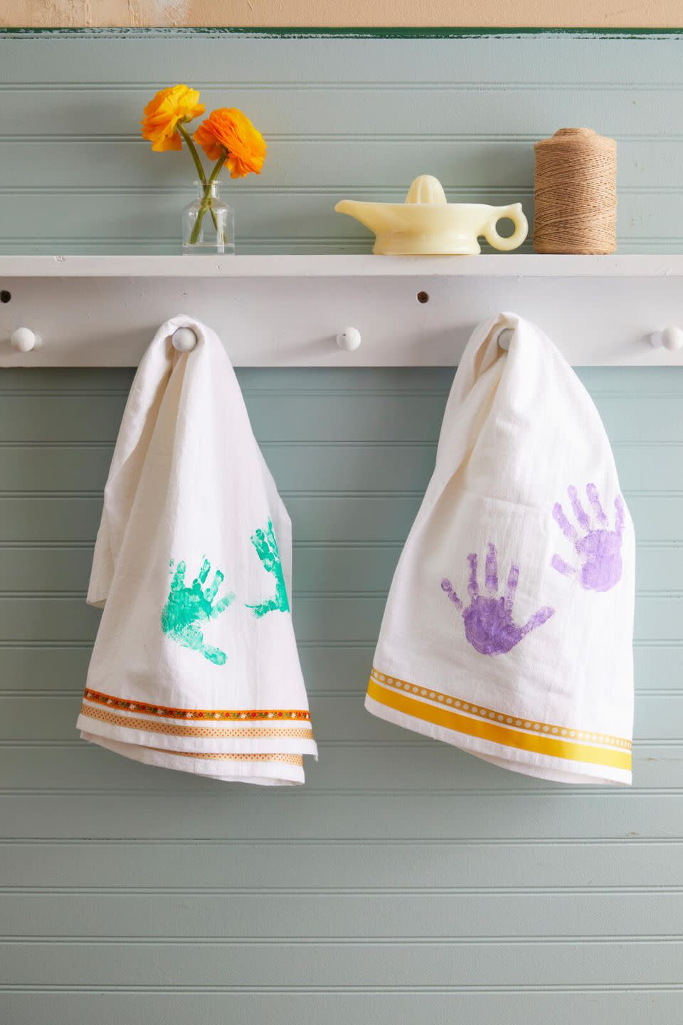 white dishtowels with ribbon sewn on the bottom edge and kids hand prints painted on the body of the towel hung from a peg rail shelf