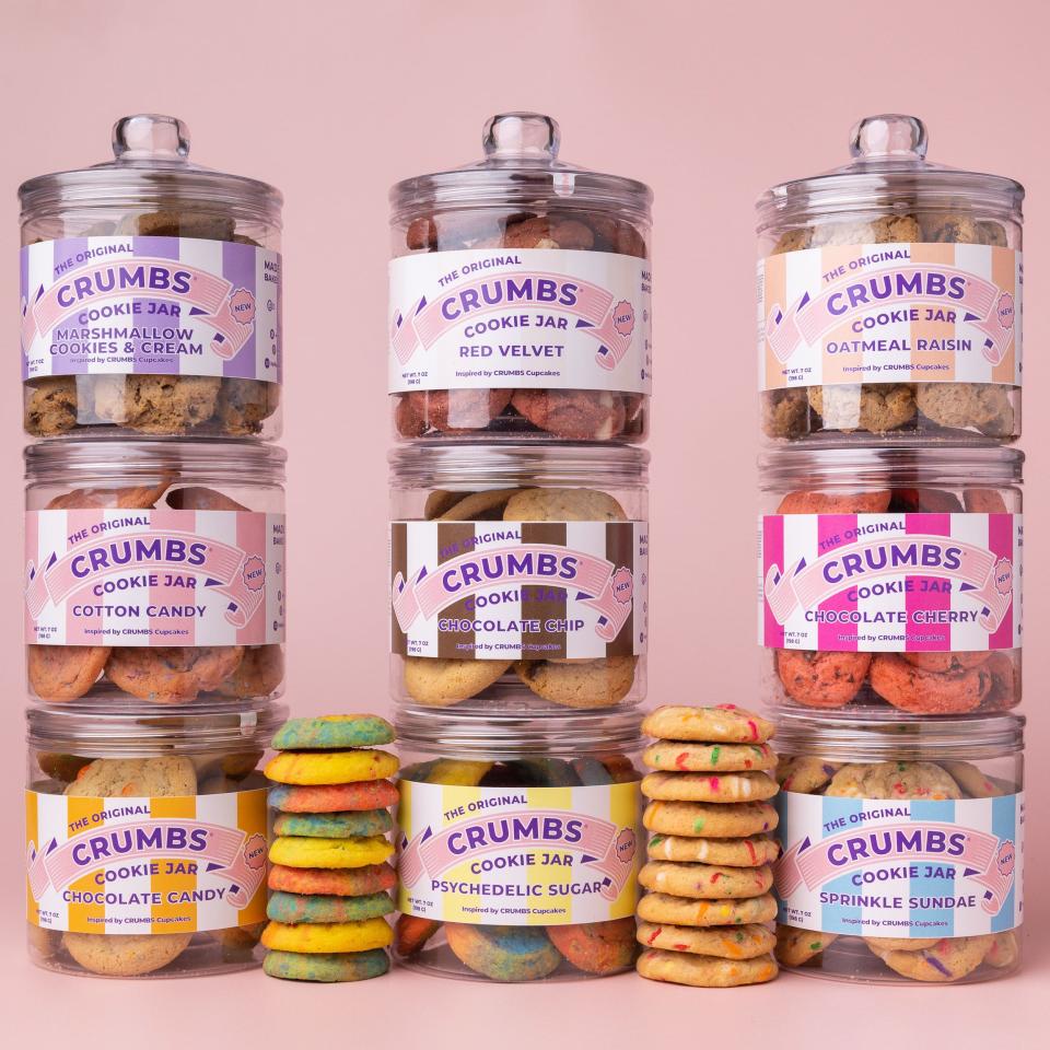 Jars of Crumbs' cookies, which come in flavors including chocolate cherry and oatmeal rasin, are stacked on top of each other against a light pink background.