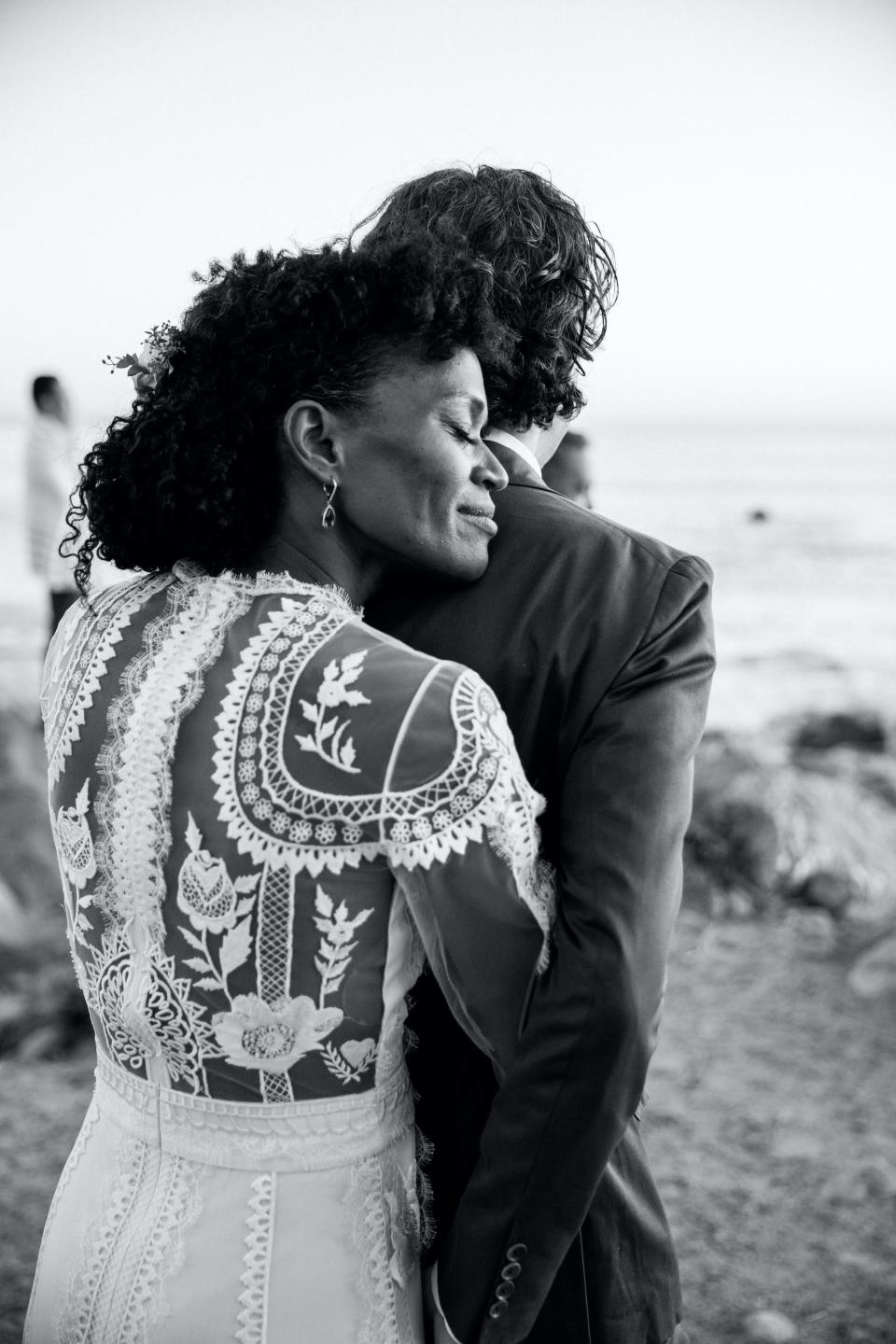 A bride hugs her groom from behind on their wedding day on a beach.