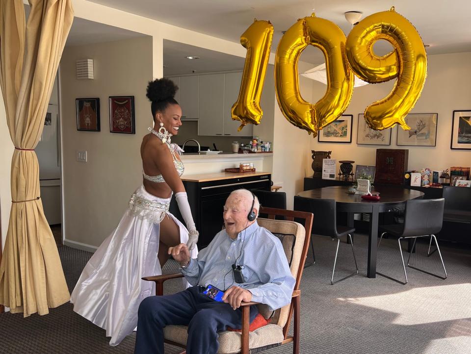 Morrie Markoff laughs under balloons reading '109' as a belly dancer goes aroudn his chair.