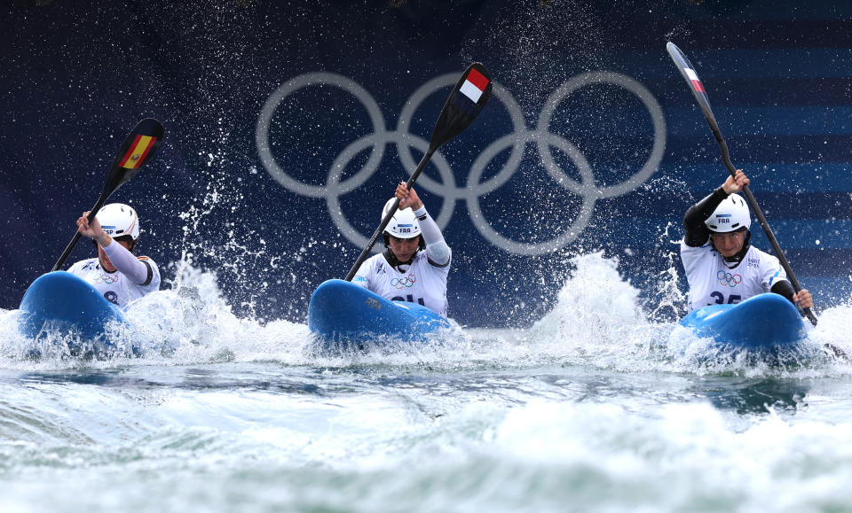 Three Olympic kayakers paddle through rough waters against a backdrop of large Olympic rings