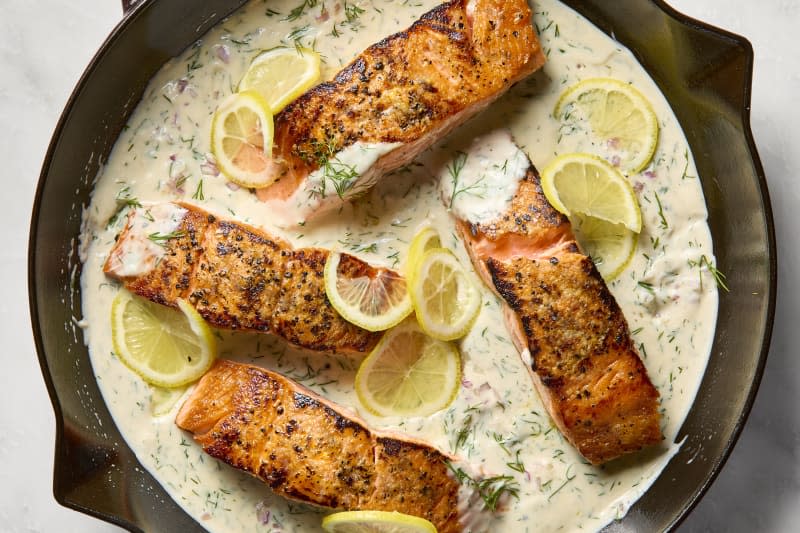 overhead shot of four pieces of salmon in a creamy lemon dill sauce, in a cast iron pan, topped with lemon wheels