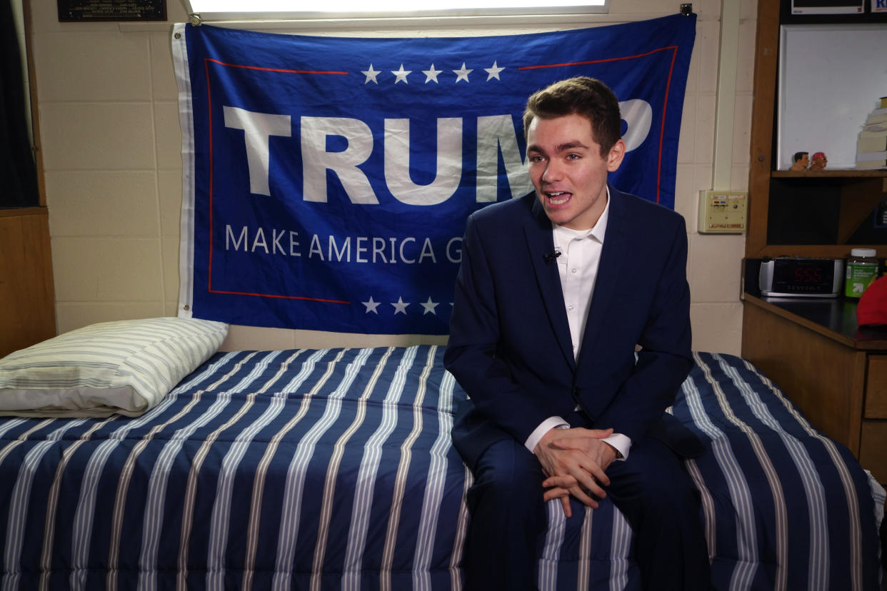 Nick Fuentes sits on a bed during an interview. Behind him is a banner supporting Donald Trump.