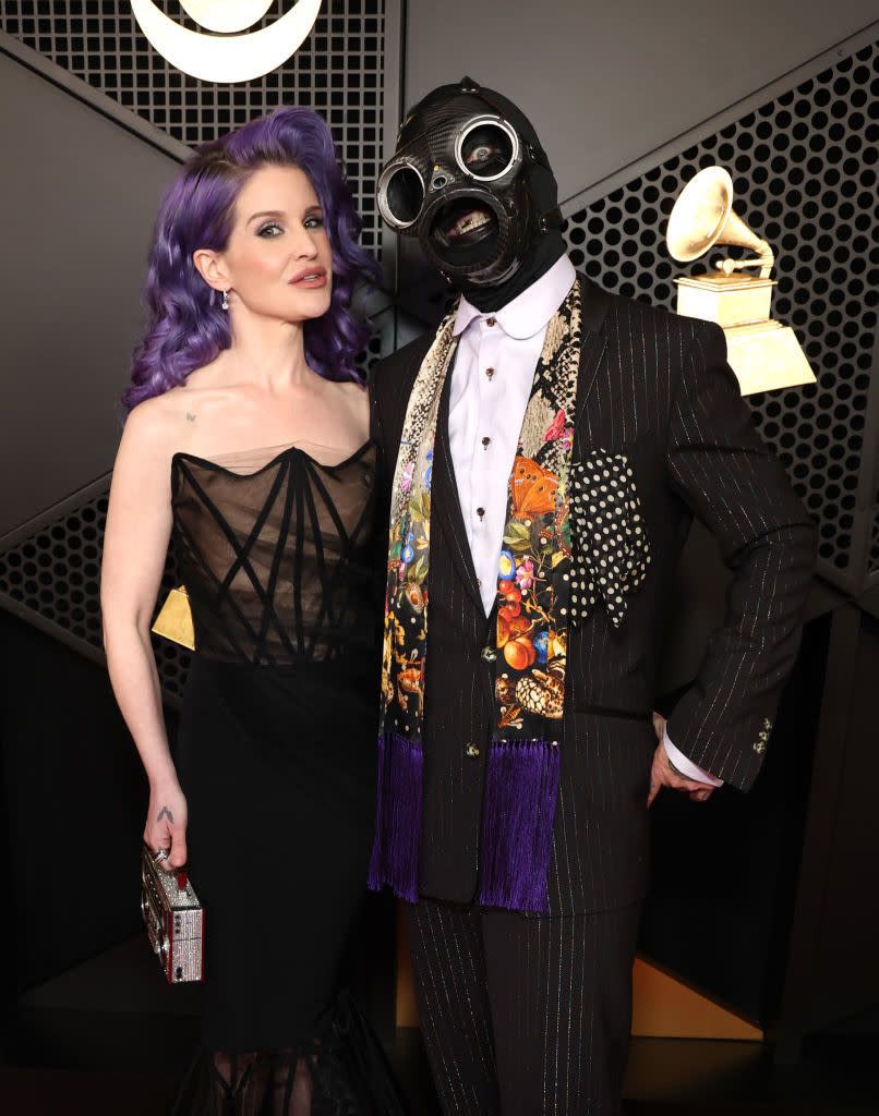 Two individuals on the red carpet. The woman is wearing a strapless black dress and holding a clutch. The man is in a suit, mask, and scarf with decorative patterns