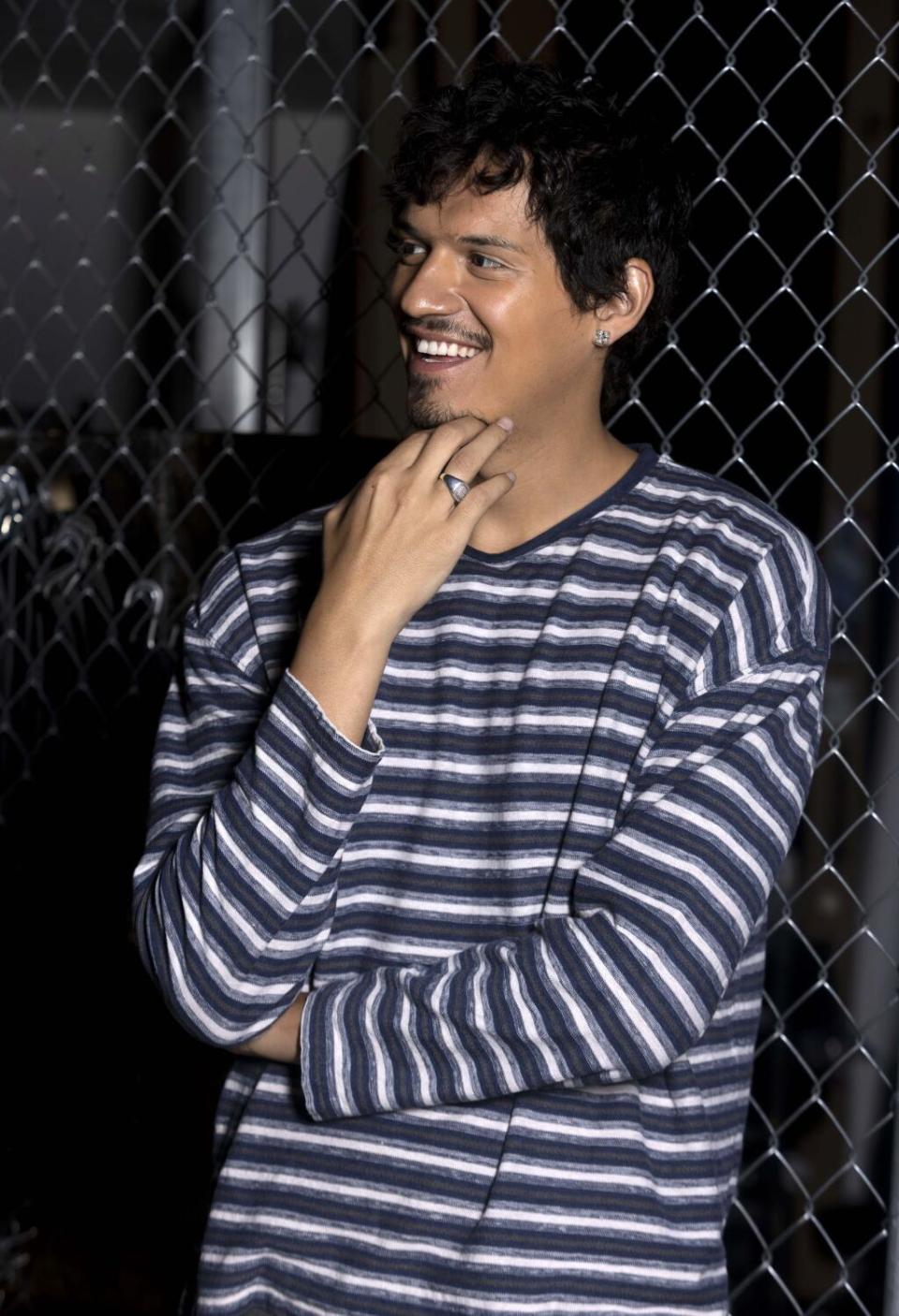 Omar Apollo wearing a striped shirt, smiling with his hand to his chin.