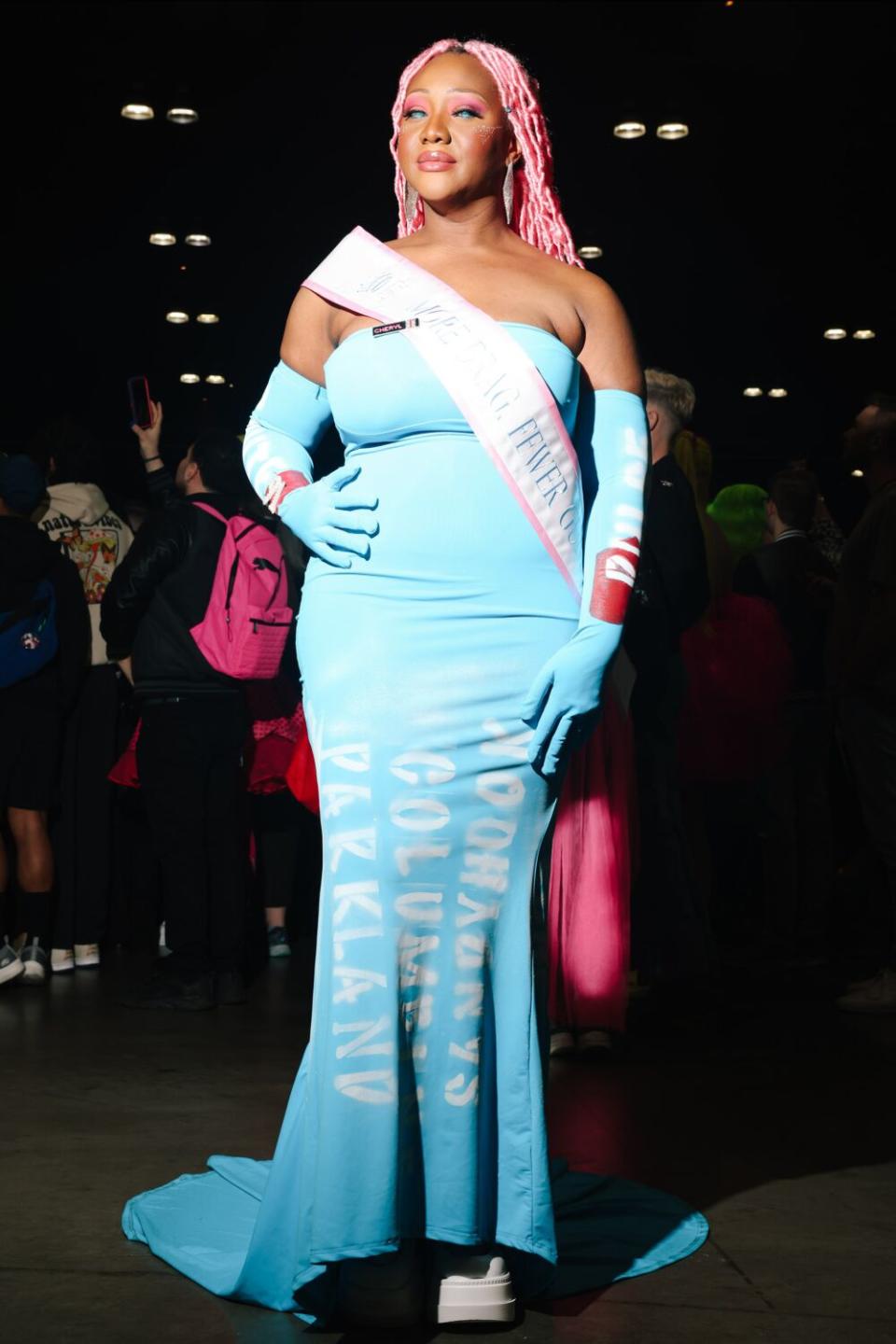 A person poses in a light blue dress and white sash.