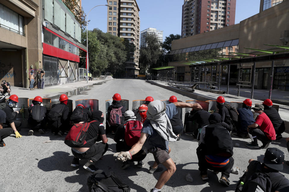 Anti-government protesters squat down behind makeshift shields made from metal barrels during clashes with police in Santiago, Chile, Thursday, Nov. 14, 2019. Students in Chile began protesting nearly a month ago over a subway fare hike. The demonstrations have morphed into a massive protest movement demanding improvements in basic services and benefits, including pensions, health, and education. (AP Photo/Esteban Felix)