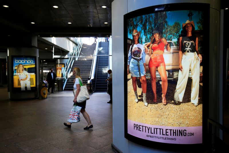 A shopper walks pass advertising billboards for Boohoo and for 'Pretty Little Things', a Boohoo brand, at Canary Wharf DLR station in central London