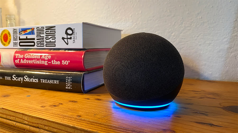 The Echo Dot smart speaker sits on a wooden shelf next to a stack of books. 