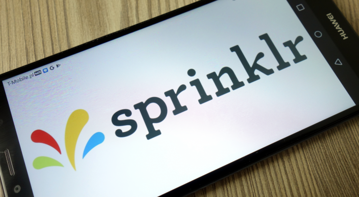 The Sprinklr (CXM) logo on a smartphone sitting on a wood table.