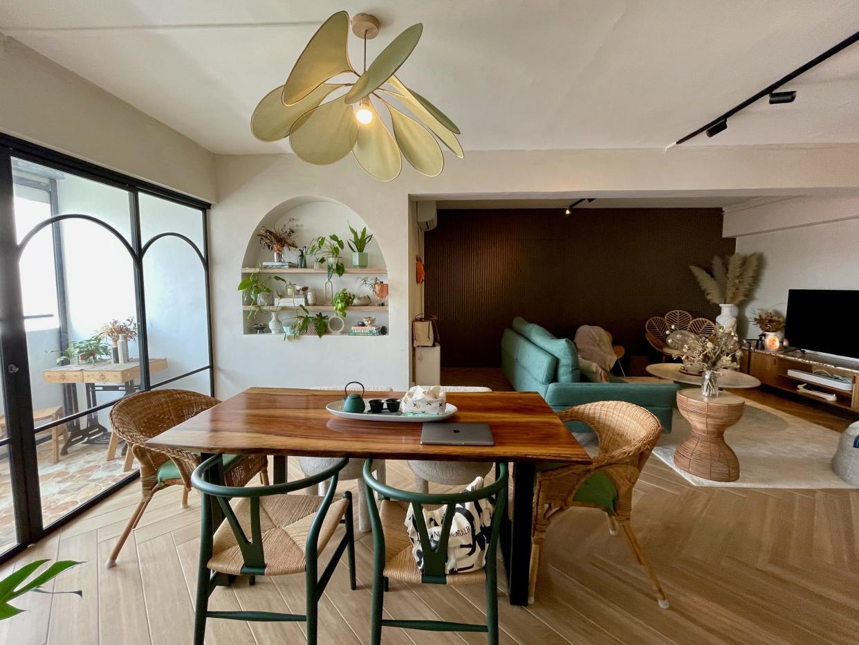 The open-plan living space in the couple's home, featuring a wooden dining table with wicker chairs, a shelf of plants, and a living space with couches and a TV off to the right..