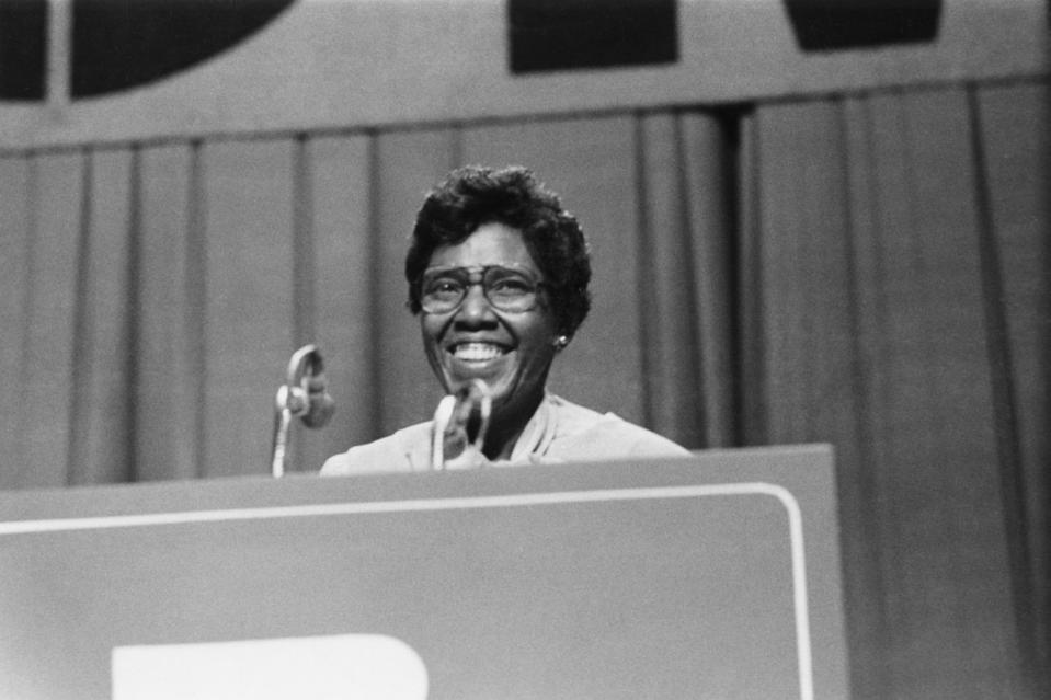 barbara jordan smiles as she stands behind a podium with a microphone, she wears glasses earrings and a light colored top